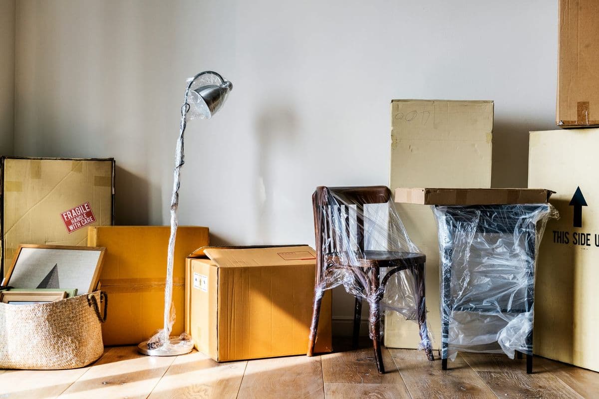 Stacked cardboard moving boxes labeled "Fragile" and "This side up" beside a shrink-wrapped chair and lamp on a hardwood floor.